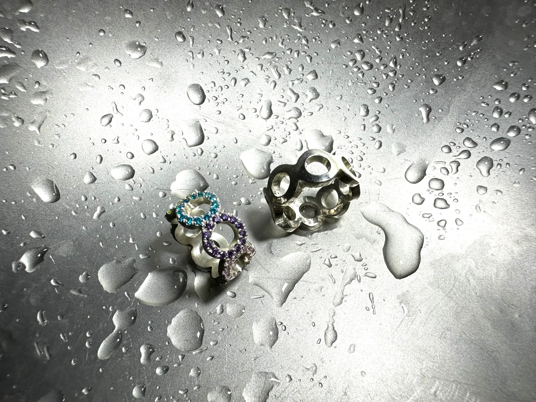 Zwei Infinity-Ringe aus Silber auf nassem Metall, einer mit blauen und violetten Saphiren besetzt