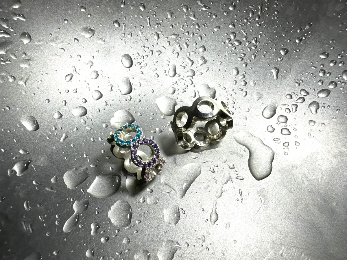 Zwei Infinity-Ringe auf nassem Metall: einer massiv in Silber, einer mit blauen und violetten Saphiren besetzt