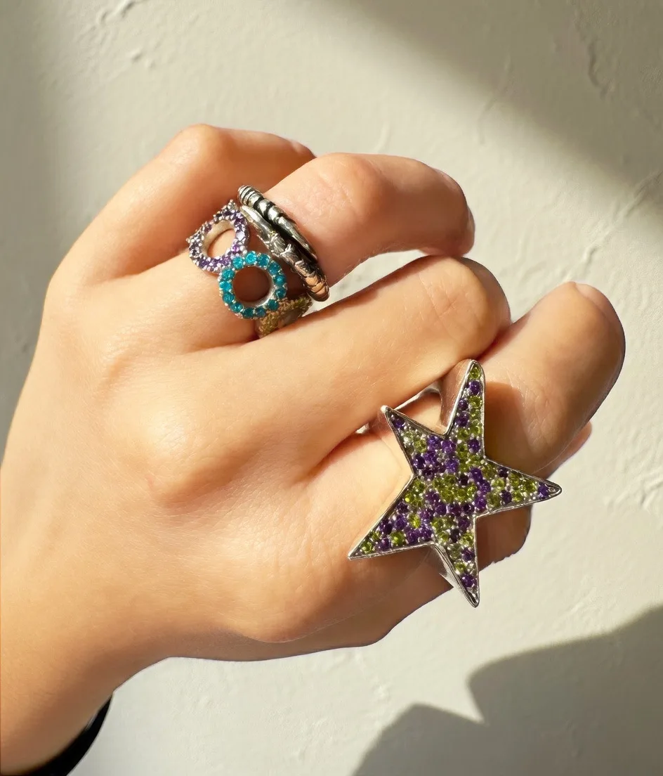 Hand mit gestapelten Silberringen und großem Sternring mit violetten und grünen Saphiren